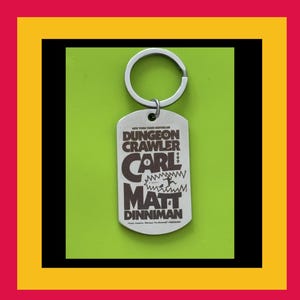May include: A silver-toned metal dog tag keyring featuring dark brown text that reads New York Times Bestseller Dungeon Crawler Carl by Matt Dinniman, with a small graphic of a figure running from a jagged mouth shape in the centre.