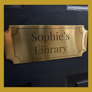 Könnte beinhalten: Rechteckige Messingplakette mit der Aufschrift "Sophie's Library" in Serifenschrift. Die Plakette ist an einer dunkelblauen Holztür mit goldenem Rand befestigt.
