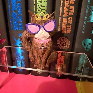 May include: A brown sculpted cat figurine wearing a gold crown and oversized purple-rimmed sunglasses with pink lenses. The cat has a pink collar with a small purple flower charm. It is positioned in front of several black books with colourful text.