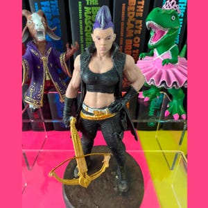 May include: A hand-painted miniature figurine of a warrior with a purple mohawk, wearing a black crop top, vest, and trousers, holding a gold crossbow. To the left is a goat-headed figure in a purple robe, and to the right is a green dinosaur in a pink tutu.