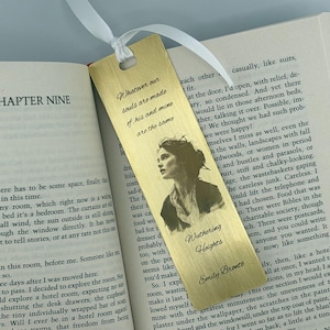 Puede incluir: Un marcapáginas de metal dorado con un retrato de mujer y texto de "Cumbres Borrascosas" de Emily Brontë. El marcapáginas está sobre un libro abierto con una cinta blanca.