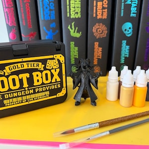 May include: A black Gold Tier Loot Box container with yellow text and graphics, a black horned creature figurine, six small paint bottles in various colours, and two paintbrushes with wooden handles, set against a row of black book spines.