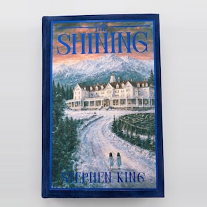 Op de afbeelding: Een hardcover boek getiteld The Shining van Stephen King met een donkerblauwe fluwelen kaft. De voorkant toont een illustratie van een groot hotel in een besneeuwd berglandschap, een doolhof en twee figuren die op een pad staan.