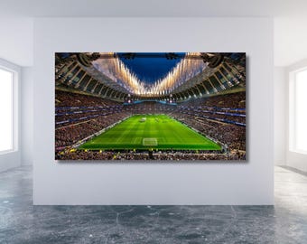 May include: A large canvas print of a football stadium at night, with a green pitch and packed stands. Fireworks explode above the stadium, creating a vibrant scene. The print is displayed on a white wall, showcasing the stadium's architecture.