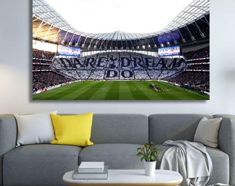 May include: A canvas print depicting a packed football stadium with the words "DARE TO DREAM" displayed by the fans. The stadium has a green pitch and a large roof. The print is mounted above a grey sofa with yellow and white cushions.