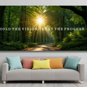 Puede incluir: Un gran lienzo que presenta una escena de bosque vibrante con un camino que conduce a un brillante estallido solar. El texto "HOLD THE VISION, TRUST THE PROCESS" se muestra en la parte superior. La impresión cuelga sobre un sofá gris con coloridas almohadas.