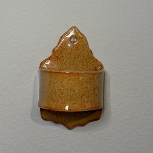 May include: A wall-mounted ceramic pocket with a speckled tan and brown glaze. The piece features a pointed top section with a single pre-drilled hole for mounting and a curved, open-front container base with a scalloped bottom edge.