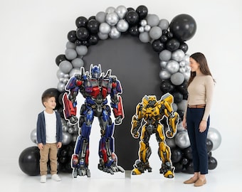 May include: Cardboard cutouts of Optimus Prime and Bumblebee stand in front of a balloon arch. The Optimus Prime cutout is red, blue, and silver. The Bumblebee cutout is yellow and black. A woman and a child are standing nearby.