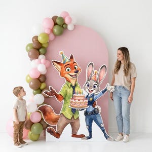 May include: A birthday party scene featuring cardboard cutouts of Nick Wilde and Judy Hopps from Zootopia, holding a cake. A young child and a woman stand nearby. The scene is decorated with a pink arch and a balloon garland.