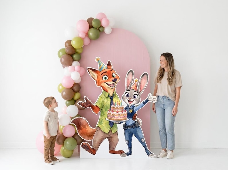 May include: A birthday party scene featuring cardboard cutouts of Nick Wilde and Judy Hopps from Zootopia, holding a cake. A young child and a woman stand nearby. The scene is decorated with a pink arch and a balloon garland.