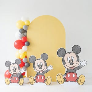 Puede incluir: Un tel&oacute;n de fondo de arco amarillo con tres recortes de Mickey Mouse frente a una guirnalda de globos. Los recortes de Mickey Mouse son negros, rojos y amarillos. La guirnalda de globos incluye globos rojos, amarillos, grises y blancos.
