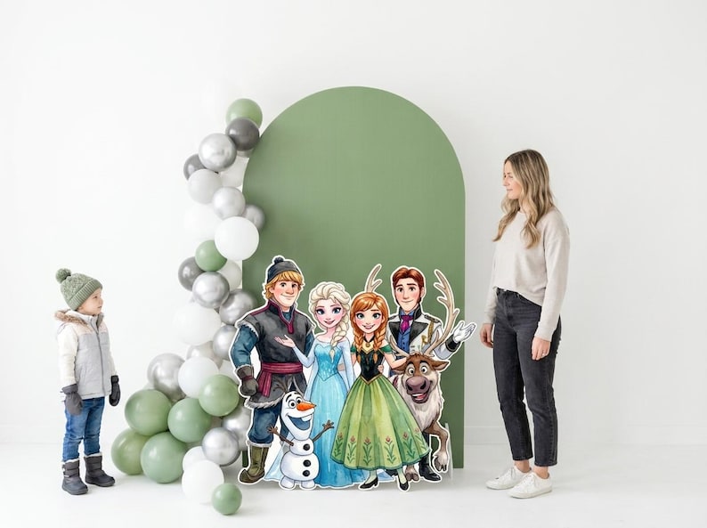 May include: A party scene featuring cardboard cutouts of characters from the animated film "Frozen", including Anna, Elsa, Kristoff, and Olaf. A young child and an adult woman are also present. A balloon arch in shades of green, silver, and white decorates the background.