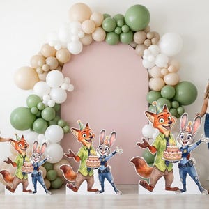 May include: Party decorations featuring Zootopia characters, including Nick Wilde and Judy Hopps, holding a cake. A balloon arch in neutral tones frames a pink backdrop. A young girl and a woman stand nearby.