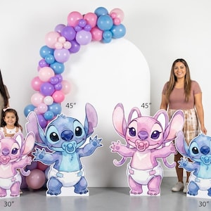 May include: Four character cutouts featuring blue and pink creatures with large ears, wearing diapers and pacifiers. Two cutouts are 45 inches tall and two are 30 inches tall. A balloon arch in shades of pink, purple, and blue is positioned behind them.