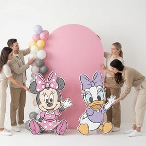 Puede incluir: Una escena de fiesta con recortes de Minnie Mouse y Daisy Duck, un gran tel&oacute;n de fondo rosa y un arco de globos. La gente est&aacute; organizando las decoraciones. Los personajes visten sus trajes caracter&iacute;sticos, con lunares y lazos.