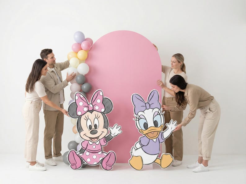Puede incluir: Una escena de fiesta con recortes de Minnie Mouse y Daisy Duck, un gran tel&oacute;n de fondo rosa y un arco de globos. La gente est&aacute; organizando las decoraciones. Los personajes visten sus trajes caracter&iacute;sticos, con lunares y lazos.
