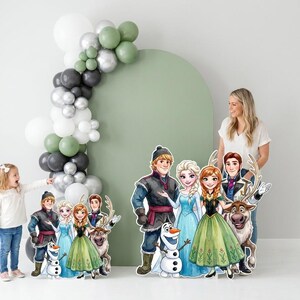 May include: A party scene featuring cardboard cutouts of characters from the animated film "Frozen." The cutouts include Anna, Elsa, Kristoff, Olaf, and Sven. A balloon arch in shades of green, gray, silver, and white decorates the background.