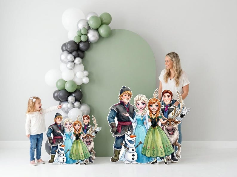May include: A party scene featuring cardboard cutouts of characters from the animated film "Frozen." The cutouts include Anna, Elsa, Kristoff, Olaf, and Sven. A balloon arch in shades of green, gray, silver, and white decorates the background.