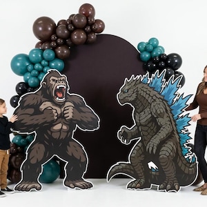 May include: Cardboard cutouts of King Kong and Godzilla stand before a dark backdrop, decorated with balloons in shades of brown, teal, and black. A woman and a child are positioned near the cutouts. The scene is set against a white background.