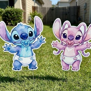 May include: Two decorative yard signs featuring cartoon characters in diapers with pacifiers. One character is blue with large ears and arms outstretched, while the other is pink with similar features. Both figures are standing on metal stakes.