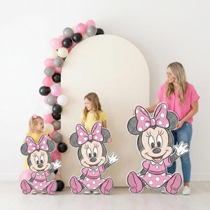 Puede incluir: Tres grandes recortes de Minnie Mouse con vestidos y lazos de lunares rosas. Una mujer y dos ni&ntilde;os sostienen los recortes. Un arco de globos con globos rosas, negros, blancos y grises est&aacute; en el fondo.