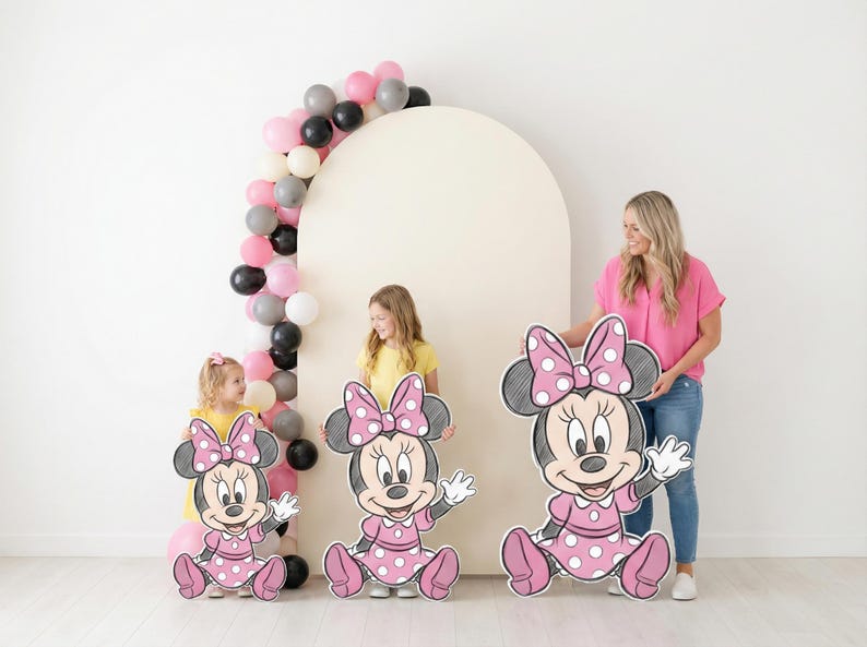 Puede incluir: Tres grandes recortes de Minnie Mouse con vestidos y lazos de lunares rosas. Una mujer y dos ni&ntilde;os sostienen los recortes. Un arco de globos con globos rosas, negros, blancos y grises est&aacute; en el fondo.
