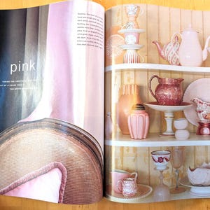 May include: A magazine spread featuring a collection of pink and peach-colored teacups, teapots, pitchers, and decorative vases displayed on white shelves. The word "pink" is visible on the left page. The overall aesthetic is vintage and elegant.