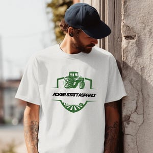 Könnte beinhalten: Weißes T-Shirt mit einem grünen Traktorgrafik und dem Text "ACKER STATT ASPHALT". Das Design beinhaltet ein Feld und ein kleines Haus. Das T-Shirt ist aus einem weichen Material.