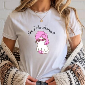 Könnte beinhalten: Weißes T-Shirt mit einer Cartoon-Katze, die eine Sonnenbrille, ein rosa Handtuch und rosa Hausschuhe trägt. Der Text "Am I the drama?" steht in handschriftlicher Schrift über der Katze. Das Shirt ist ein lässiger Stil.