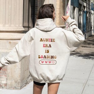 Könnte beinhalten: Cremefarbener Kapuzenpullover mit dem Text "AUNTIE ERA IS LOADING" in braunen, rosa und braunen Buchstaben. Darunter befinden sich drei rosa Herzen in einem braunen Rechteck. Der Hoodie hat eine Kapuze und lange Ärmel.