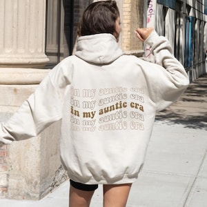 Könnte beinhalten: Beigefarbener Kapuzenpullover mit dem Schriftzug "in my auntie era" in wellenförmiger, brauner Schrift auf dem Rücken. Der Hoodie hat eine Kapuze und lange Ärmel. Die Person trägt schwarze Shorts.