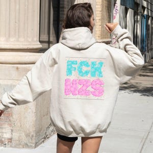 May include: A beige hoodie with a hood and long sleeves. The back of the hoodie features the text "FCK NZS" in a blue and pink bubble letter design. The hoodie is made of a soft material.