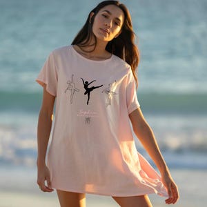 Könnte beinhalten: Hellrosa T-Shirt mit drei Strichzeichnungen von Ballerinas. Darunter steht der Text "Sophia ROSE O DANCE". Das kurzärmlige T-Shirt hat eine lockere Passform und ist für den Alltag geeignet.