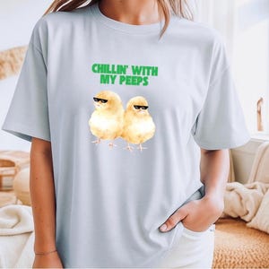 Könnte beinhalten: Hellblaues T-Shirt mit dem grünen Text "CHILLIN' WITH MY PEEPS" und einer Grafik von zwei gelben Küken mit Sonnenbrille. Das Shirt besteht aus einem weichen Material.