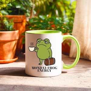 Könnte beinhalten: Ein weißer Keramikbecher mit hellgrünem Innenraum und Henkel, der eine Grafik eines grünen Frosches zeigt, der eine kleine weiße Kaffeetasse und einen braunen Koffer hält, mit dem schwarzen Text MONDAY FROG ENERGY unter der Abbildung.