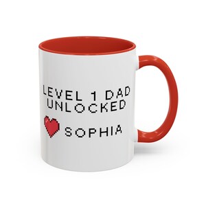 May include: White ceramic mug with a red interior and handle. The mug features the text "LEVEL 1 DAD UNLOCKED SOPHIA" in black pixelated font, along with a red pixelated heart graphic. A fun gift for a new dad.