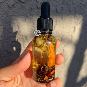 May include: A clear glass bottle with a black dropper top, filled with a golden-hued liquid and botanical ingredients. The bottle has a silver decorative ring. The contents include dried herbs and flowers, creating a natural aesthetic.