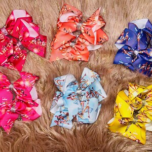 May include: Six colorful hair bows in shades of red, orange, blue, pink, light blue, and yellow. Each bow features a rhinestone center and a printed design with text and figures. The bows are arranged on a fluffy, brown surface.