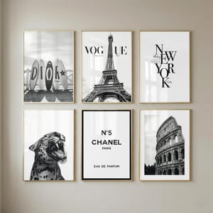 May include: A set of six black and white wall art prints in gold frames. The prints feature surfboards with the text DIOR, the Eiffel Tower with VOGUE, the text NEW YORK City, a leopard, a Chanel N°5 perfume label, and the Roman Colosseum.