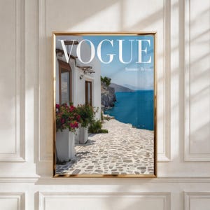 Puede incluir: Una impresión de arte mural enmarcada que muestra una escena costera mediterránea con edificios blancos, buganvillas rosas, un camino de piedra y un mar azul profundo. La palabra VOGUE está impresa en letras blancas grandes en la parte superior.
