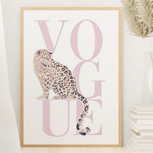 Könnte beinhalten: Gerahmter Druck mit einer Aquarell-Illustration eines Leoparden und dem Wort "VOGUE" in Rosa. Der Leopard hat ein geflecktes Fell und ist vor den Buchstaben positioniert. Der Rahmen hat eine helle Holzfarbe.