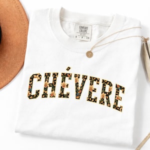 Puede incluir: Una camiseta blanca con la palabra CHÉVERE impresa en una fuente arqueada y llamativa. Las letras están rellenas con un patrón botánico oscuro de flores amarillas y pájaros pequeños. La camiseta tiene cuello redondo y una etiqueta Comfort Colors.
