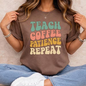 Puede incluir: Una camiseta marrón con el texto "TEACH COFFEE PATIENCE REPEAT" en letras de estilo retro. El texto es en tonos turquesa, coral, amarillo y beige. La camiseta se combina con vaqueros azules y zapatillas blancas.