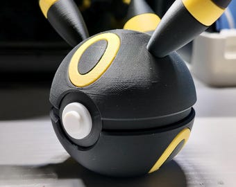 May include: Black and yellow 3D-printed Umbreon-themed Poke Ball. The ball features a circular design with yellow accents and a white button. The design includes two black, pointed ears with yellow tips.
