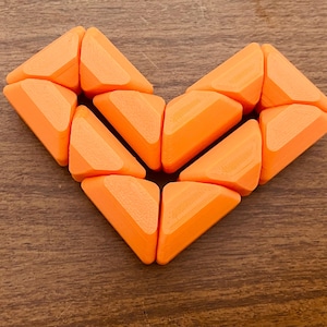 Può includere: Una composizione a forma di cuore composta da dieci pezzi di puzzle triangolari in plastica di colore arancione brillante. Ogni pezzo presenta una finitura opaca liscia con linee di strato stampate in 3D visibili, creando un cuore geometrico.