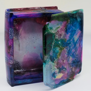 May include: A colorful, open book-shaped box with a lid. The box is a mix of purple, blue, and green hues with glitter accents. The interior is visible, showing a similar color palette. The box is likely a decorative item or a small storage container.