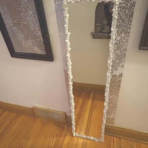 Puede incluir: Un espejo alto y rectangular con un marco de cuentas. El marco está adornado con pequeñas cuentas blancas y acentos más grandes con forma de perla. El espejo está apoyado contra una pared con papel pintado estampado y suelo de madera.