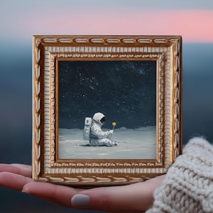 May include: A framed painting depicts an astronaut seated on a lunar surface, holding a yellow flower. The artwork is set within a gold-colored, ornate frame. The background shows a starry night sky.