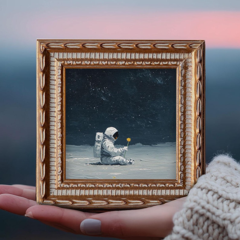 May include: A framed painting depicts an astronaut seated on a lunar surface, holding a yellow flower. The artwork is set within a gold-colored, ornate frame. The background shows a starry night sky.