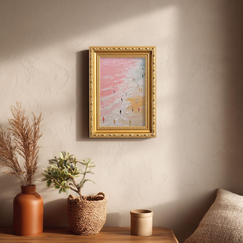 May include: A framed oil painting depicts a beach scene with a pink sky and yellow sand. The artwork features small figures of people walking along the shore. The gold frame adds a classic touch to the artwork.